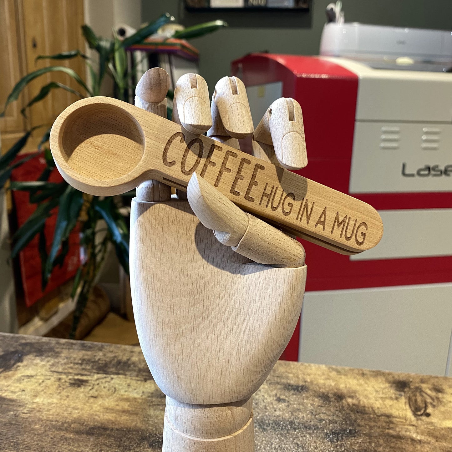 A wooden artist hand holding a coffee spoon engraved with "Hug in a Mug," set against a backdrop of coffee beans in a cozy studio setting.