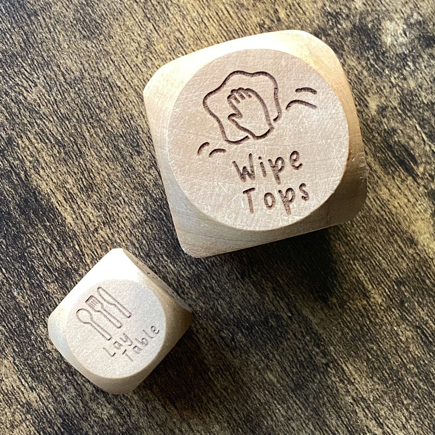 Top-down view of two sizes of Junior Chores Decision Dice, highlighting engraved chore images and natural wood grain