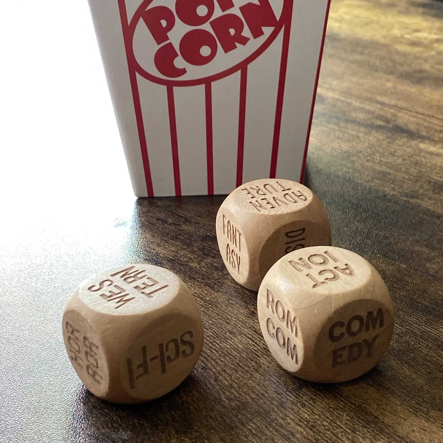 Angled shot of three wooden Movie Night Decision Dice positioned in front of a classic striped popcorn box, with engraved film genres clearly visible,