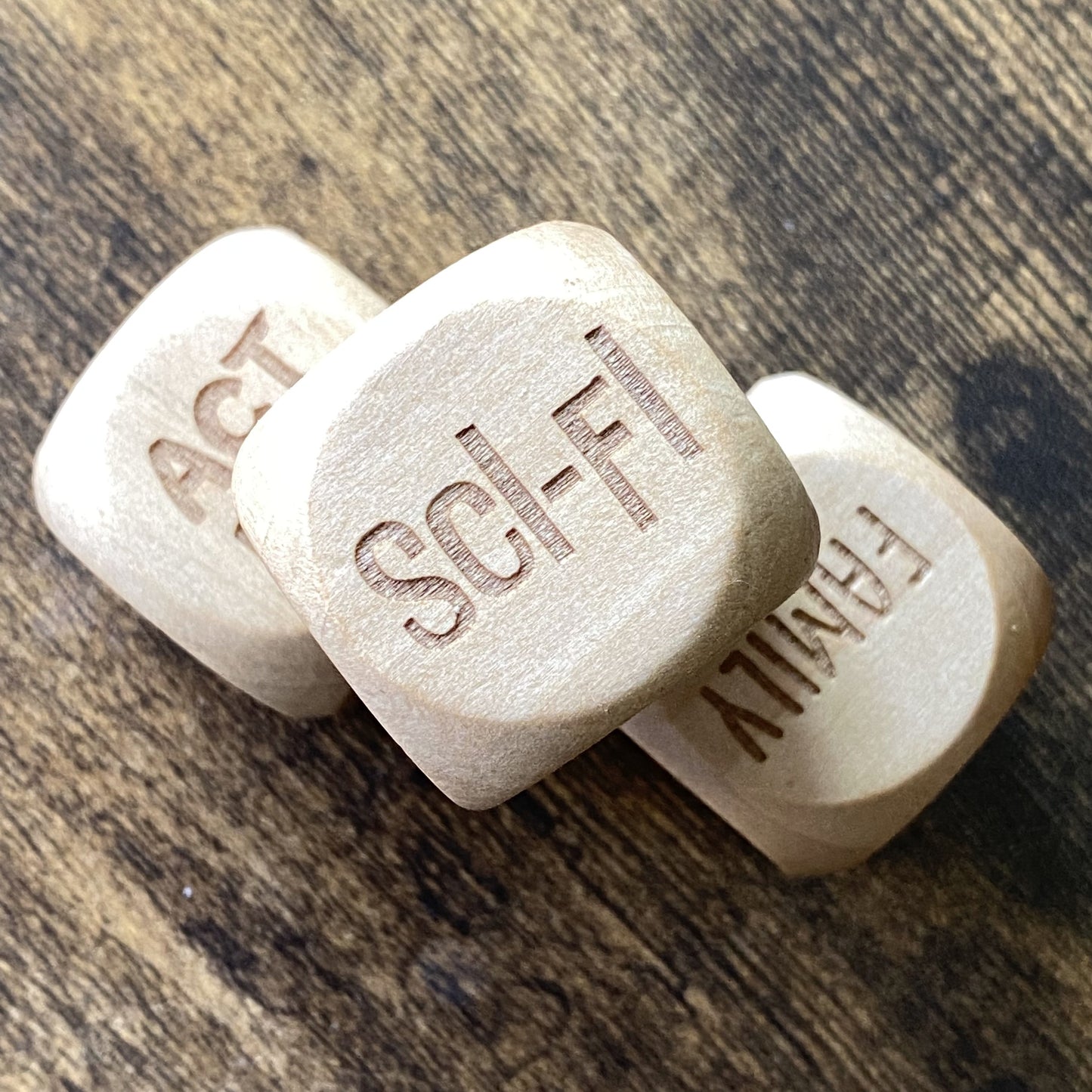 Top-down view of three wooden Movie Night Decision Dice arranged neatly on a wooden surface, showing engraved film genre options on each face for a home movie sessions