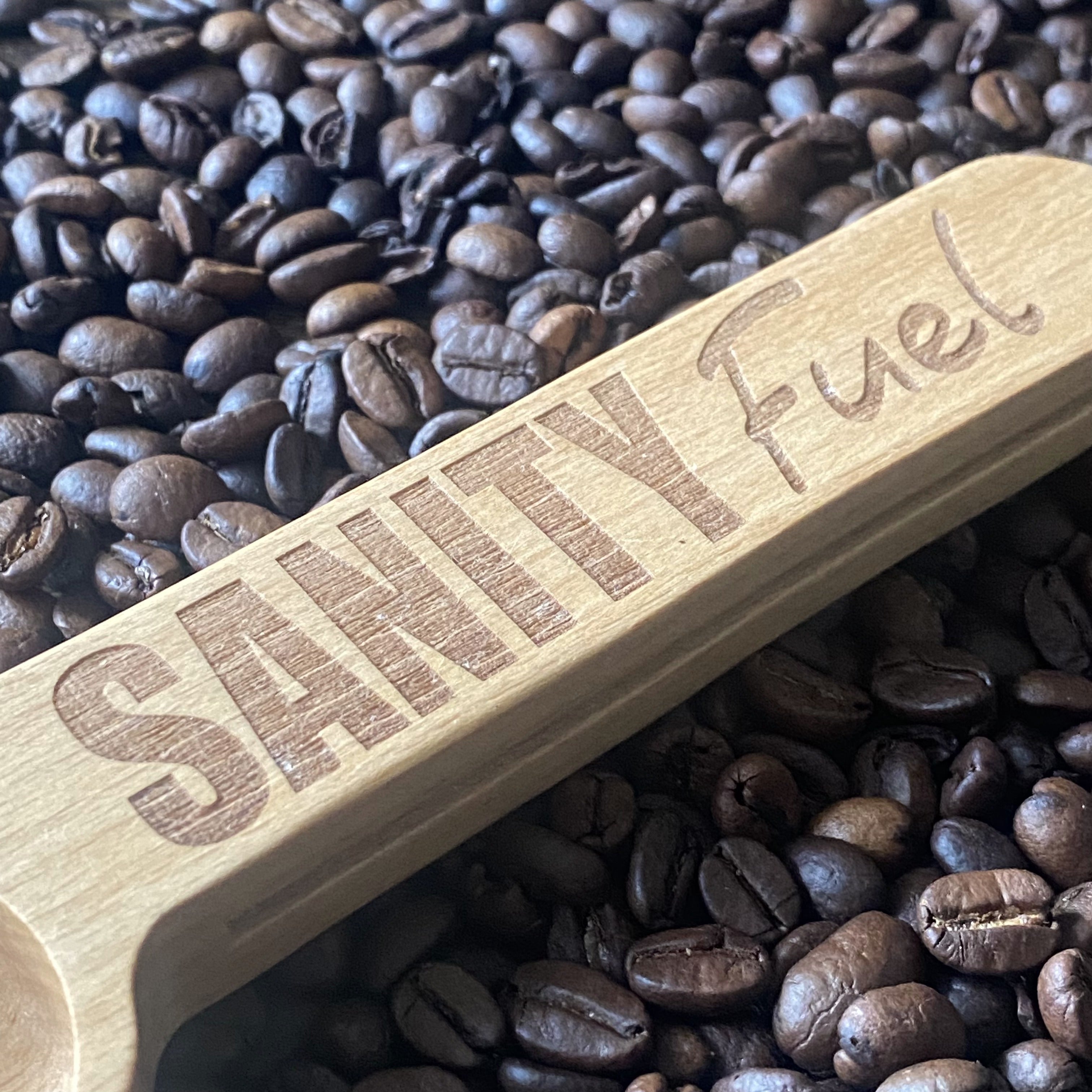 Close-up of the wooden coffee scoop resting on a bed of coffee beans, highlighting the engraved details and smooth finish.