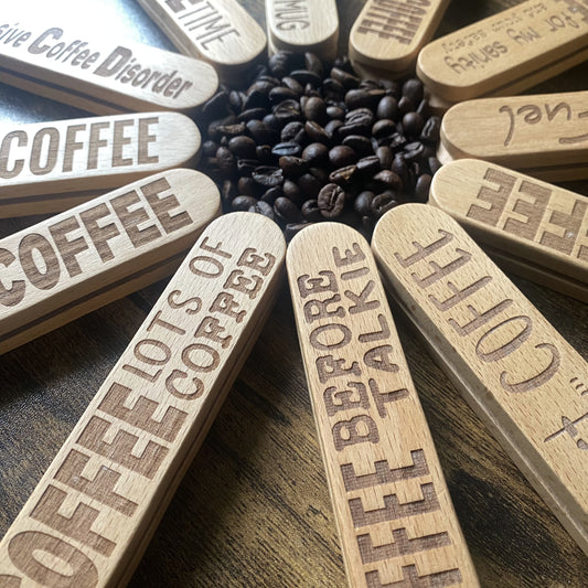 Wooden coffee scoops arranged with handles pointing inward, featuring engraved quotes, with coffee beans in the centre.