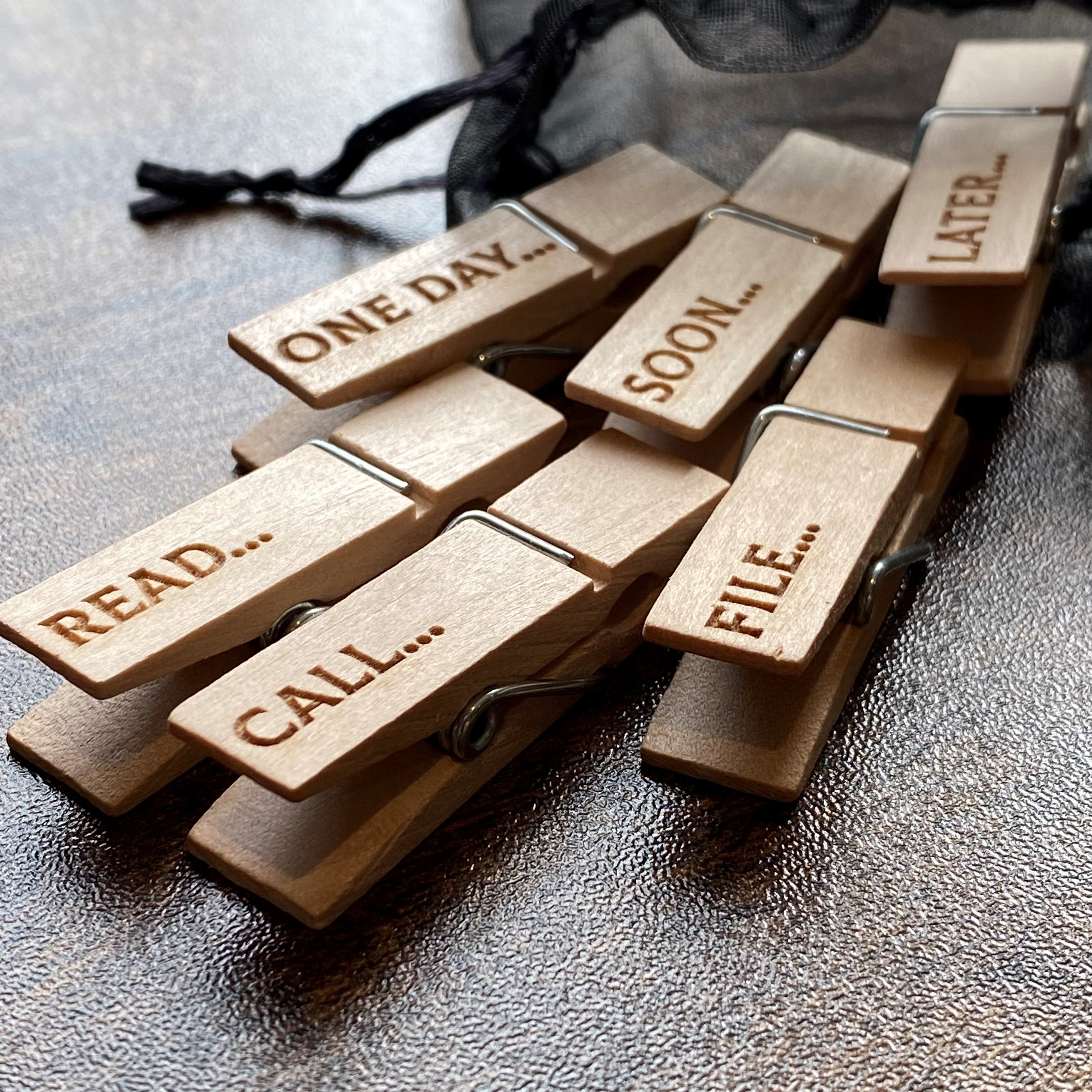 Six natural wood mini desk organisation pegs scattered from a soft organza bag on a wooden surface, showing charming engraved designs and tidy workspace vibes.