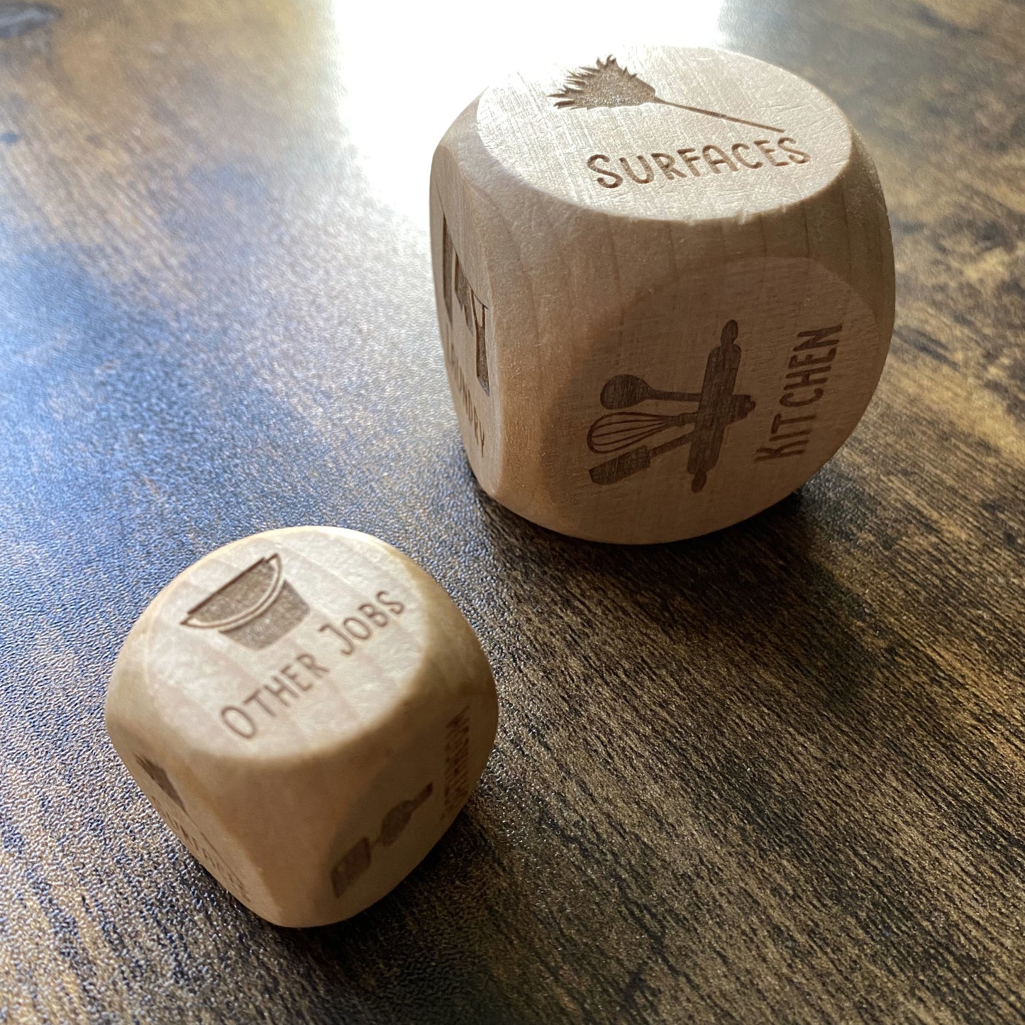 Two sizes of engraved wooden household chores dice displayed side by side, showing intricate detail engravings and natural wood texture

