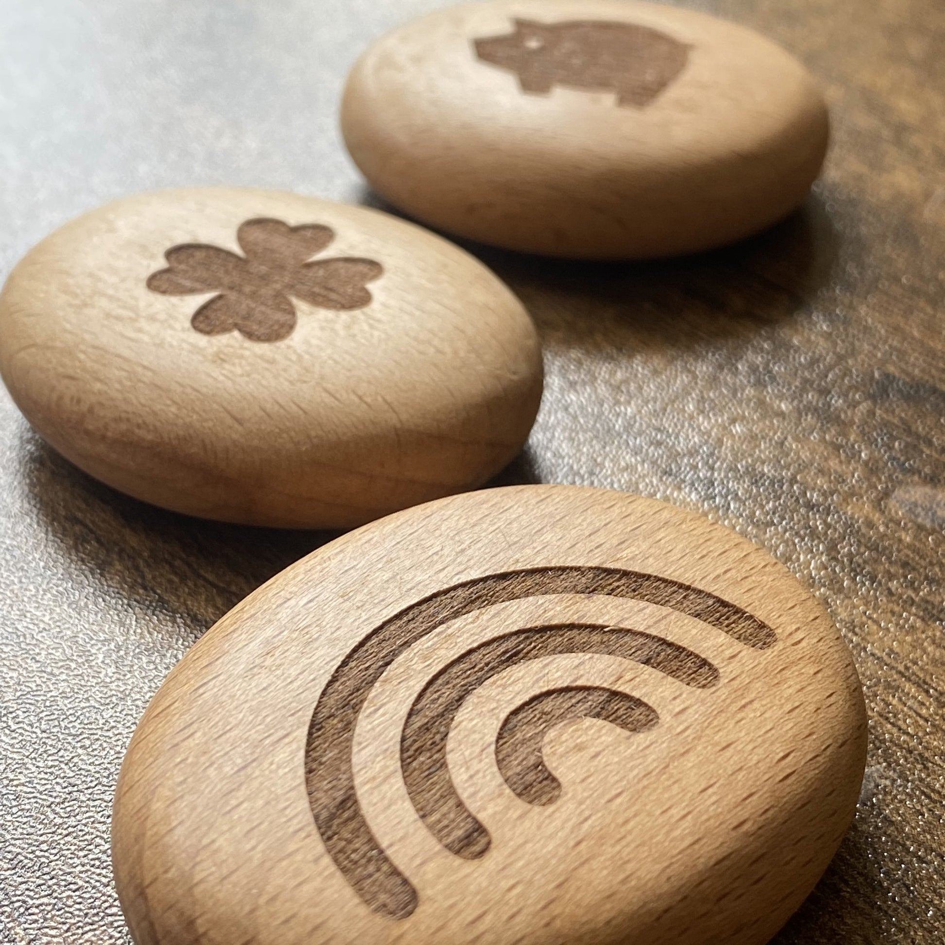 Side-angled shot of three engraved wooden pocket pebbles from the Luck Edition – featuring rainbow, clover, and lucky pig designs