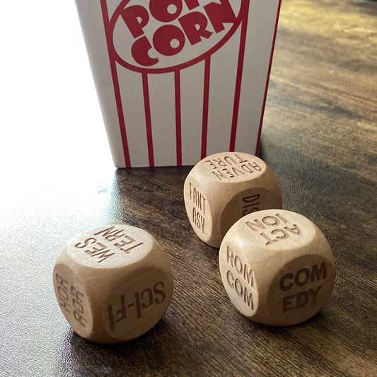 Angled shot of three wooden Movie Night Decision Dice positioned in front of a classic striped popcorn box, with engraved film genres clearly visible,