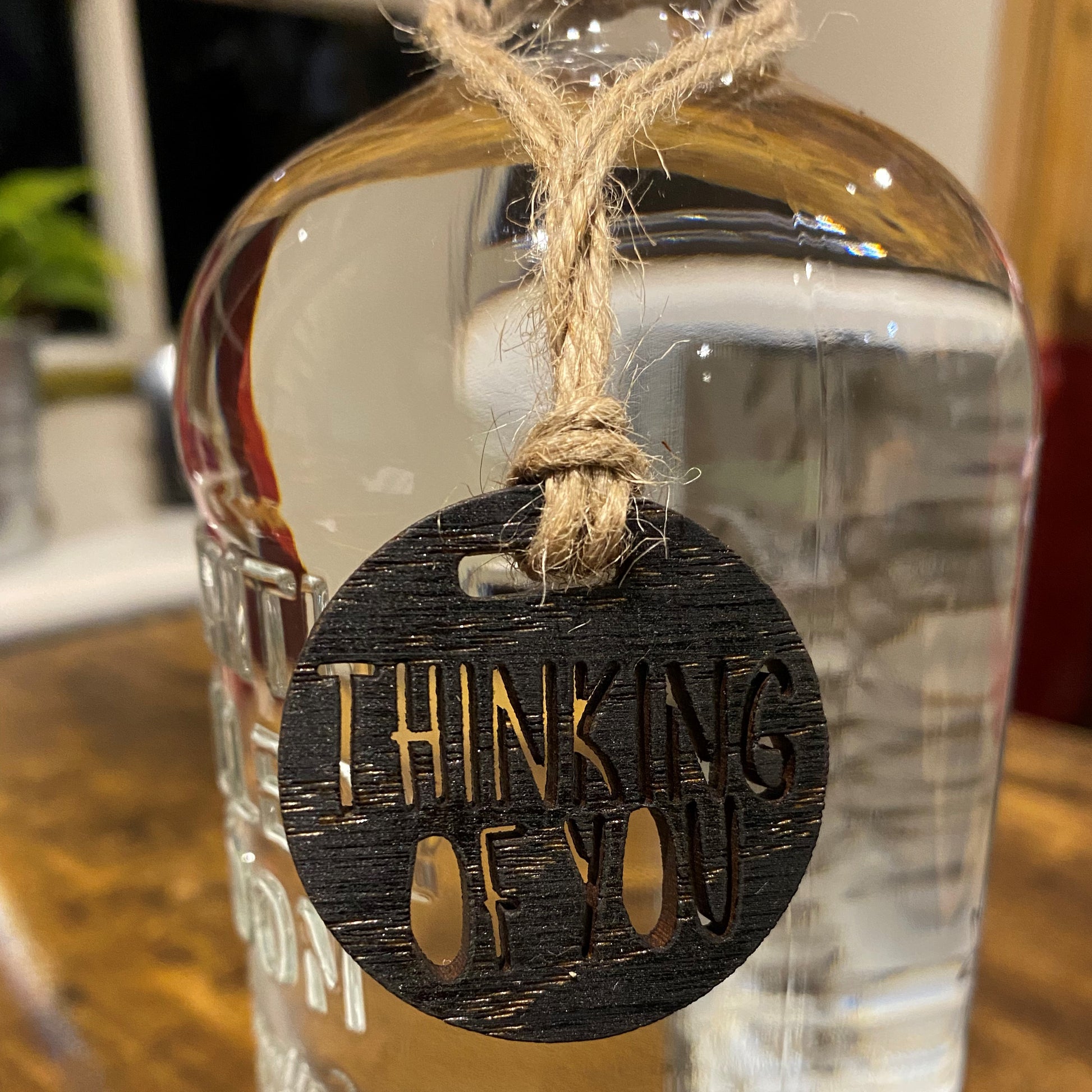 Close-up of an engraved wooden charm tied to the neck of a bottle, showcasing 'Thinking of You' design