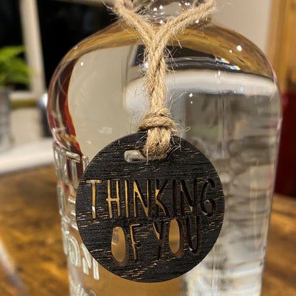 Close-up of an engraved wooden charm tied to the neck of a bottle, showcasing 'Thinking of You' design
